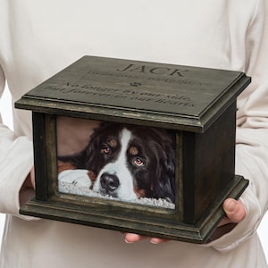 Może przedstawiać: Drewniana urna dla zwierząt w kolorze ciemnoszarym ze zdjęciem berneńskiego psa pasterskiego w ramce. Na pokrywie wygrawerowano imię "JACK", daty 29.10.2015 - 12.03.2025 oraz napis "Już nie po naszej stronie, ale na zawsze w naszych sercach."