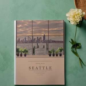 May include: A book titled "Grow Wild in Seattle" with a window illustration of the Seattle skyline. The book is on a teal surface, with a straw hat and a white flower. Small potted plants are on the window ledge.