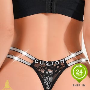 Könnte beinhalten: Schwarzer Spitzen-String mit Strass-Trägern und dem Wort "CUSTOM" auf der Vorderseite. Die Unterwäsche hat ein florales Spitzendesign und ist mit einem schwarzen BH kombiniert. Ein grüner Kreis mit dem Text "24 HOURS" und "SHIP IN" ist sichtbar.