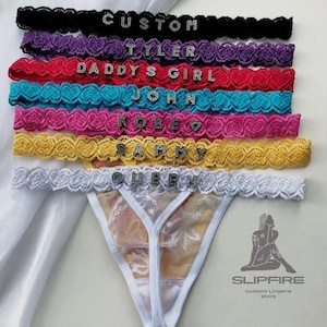 May include: A collection of custom lace garters in various colors, including black, purple, red, turquoise, pink, yellow, and white. Each garter features a different name or word spelled out in sparkling letters. A white and floral thong is also visible.