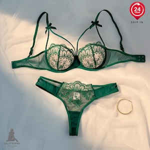 May include: A green lingerie set featuring a bra and matching thong. The bra has intricate lace detailing on the cups and delicate straps with small bows. The thong also has lace accents. A gold hoop earring is also visible.