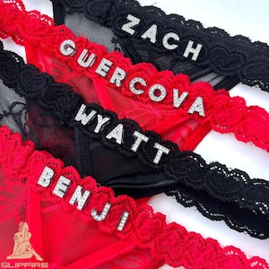 May include: Close-up of four pieces of lingerie in red and black. Each piece has a name spelled out in silver letters. The lingerie has lace trim and the names are Zach, Guercova, Wyatt, and Benji.