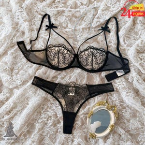 May include: Black lingerie set featuring a sheer bra with lace detailing and a matching thong. The bra has delicate bow accents on the straps. The set is displayed on a white lace fabric.
