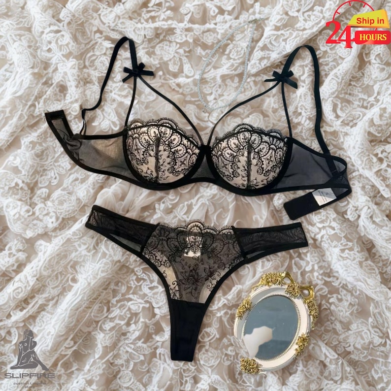May include: Black lingerie set featuring a sheer bra with lace detailing and a matching thong. The bra has delicate bow accents on the straps. The set is displayed on a white lace fabric.