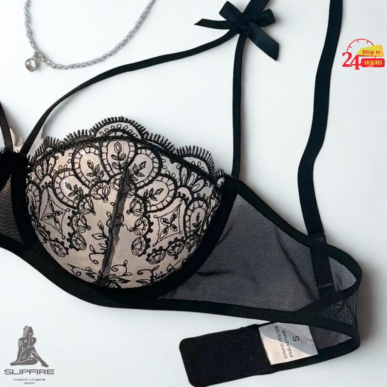 May include: Black and blush-colored bra with intricate lace detailing. The bra features adjustable straps, a delicate bow, and a sheer mesh design. A silver necklace with a pendant is also visible.