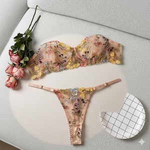Puede incluir: Un conjunto de lencería color melocotón con bordado floral. El conjunto incluye un sujetador sin tirantes y un tanga, ambos adornados con flores coloridas. También hay un ramo de rosas rosas y un plato decorativo.