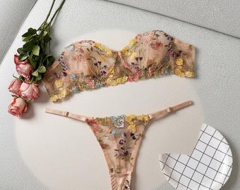 Conjunto de lingerie elegante com bordado floral, composto por sutiã e calcinha fio dental em tule transparente. Romântico, leve e perfeito para um presente íntimo.