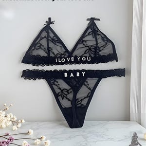 May include: Black lingerie set featuring a triangle bra and thong. The bra and thong are adorned with lace detailing and the words "I LOVE YOU" and "BABY" in white lettering. The set is displayed against a white background.