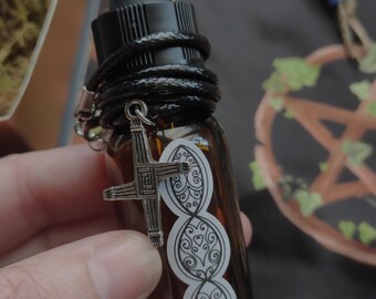May include: A brown glass essential oil bottle with a black dropper and a black leather cord. A silver-colored cross charm hangs from the cord. The bottle has three white oval stickers with decorative designs.