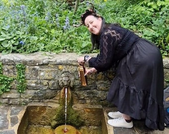 May include: A woman filling a copper-colored water bottle from a stone fountain with a lion's head spout. The fountain is surrounded by lush greenery and stone walls. The woman is wearing a black outfit and white sneakers.