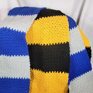 May include: A handmade crochet sweater featuring a color block design. The sweater has stripes of blue, gray, black, and yellow. The sleeves are blue and gray striped. The sweater is made of a soft, textured yarn.