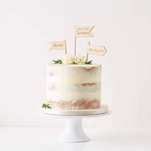May include: A white cake with a simple design, decorated with three wooden flags. The flags say "Mr. & Mrs. Simpson", "Always", and "17th June 2017".