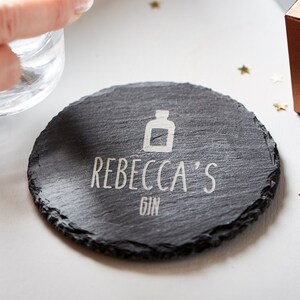 May include: A round black slate coaster with the text "Rebecca's Gin" and a small bottle icon engraved in white.