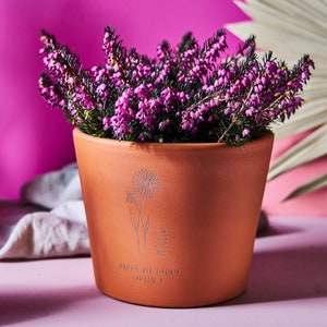 May include: A terracotta plant pot filled with purple heather. The pot features an etched floral design and the words "Happy Birthday lovely x" and the name "Milly". The background is a bright pink.