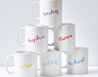 Op de afbeelding: Zes witte keramische mokken met verschillende namen erop geschreven in kleurrijke letters. De namen zijn Becky, Sophie, Rosie, Alex, Molly en Michael.
