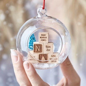 Peut inclure: Une boule de Noël en verre contenant un mini sapin de Noël en blocs de bois. Les blocs sont gravés avec "Noah Winter Jones", "N", "6lbs 2oz", "1er Noël", et "12 novembre 2018".