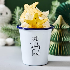 Pode incluir: Uma caneca de esmalte branco com borda azul, cheia de lanches e coberta com tortilhas em forma de estrela. A caneca tem o texto "Jacks Treats" em escrita preta. Apresentação festiva de comida.