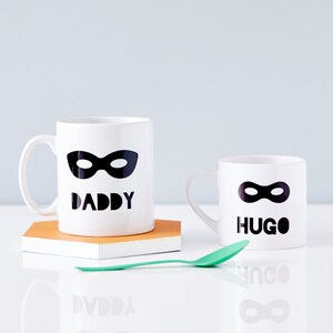 Peut inclure: Deux tasses en c&eacute;ramique blanche avec des masques de super-h&eacute;ros noirs et du texte. La plus grande tasse dit "Papa" et la plus petite tasse dit "Hugo".