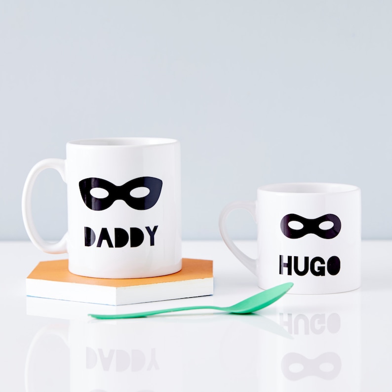 Peut inclure: Deux tasses en c&eacute;ramique blanche avec des masques de super-h&eacute;ros noirs et du texte. La plus grande tasse dit "Papa" et la plus petite tasse dit "Hugo".