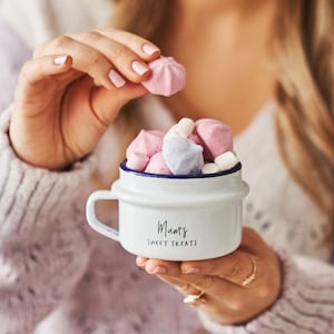 Peut inclure: Une tasse en émail blanc avec un bord et une poignée bleus. La tasse porte l'inscription "Mum's Sweet Treats" imprimée sur le devant. La tasse est remplie de guimauves roses, blanches et bleues.