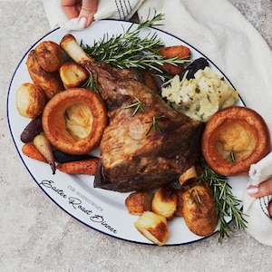 May include: A white oval platter with a blue rim, filled with a roasted leg of lamb, Yorkshire puddings, roasted potatoes, carrots, and rosemary sprigs. The platter reads "Easter Roast Dinner".