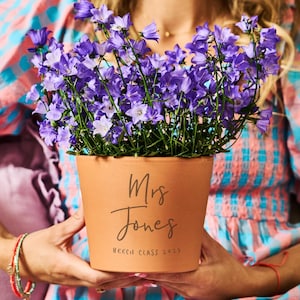 Puede incluir: Una maceta de terracota llena de campanillas moradas. La maceta está inscrita con "Mrs Jones" y "BEECH CLASS 2025". La maceta es sostenida por una persona que lleva una blusa estampada colorida.