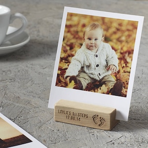 Op de afbeelding: Een houten fotostandaard met de tekst "LAYLA'S 1st STEPS 12.08.14" en een voetafdruk symbool. Een foto van een baby die in een hoop bladeren zit, wordt in de standaard weergegeven.