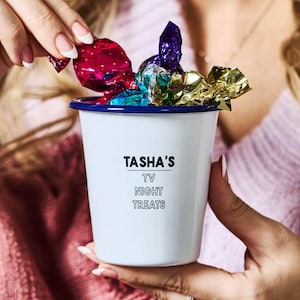 Peut inclure: Mug en émail blanc avec un bord bleu, rempli de bonbons emballés colorés. Le mug porte l'inscription "TASHA'S TV NIGHT TREATS". Le mug est tenu par une personne.