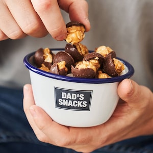 Pode incluir: Uma tigela de esmalte branco com uma borda azul e o texto "DAD'S SNACKS" impresso na frente. A tigela está cheia de pipoca coberta de chocolate.