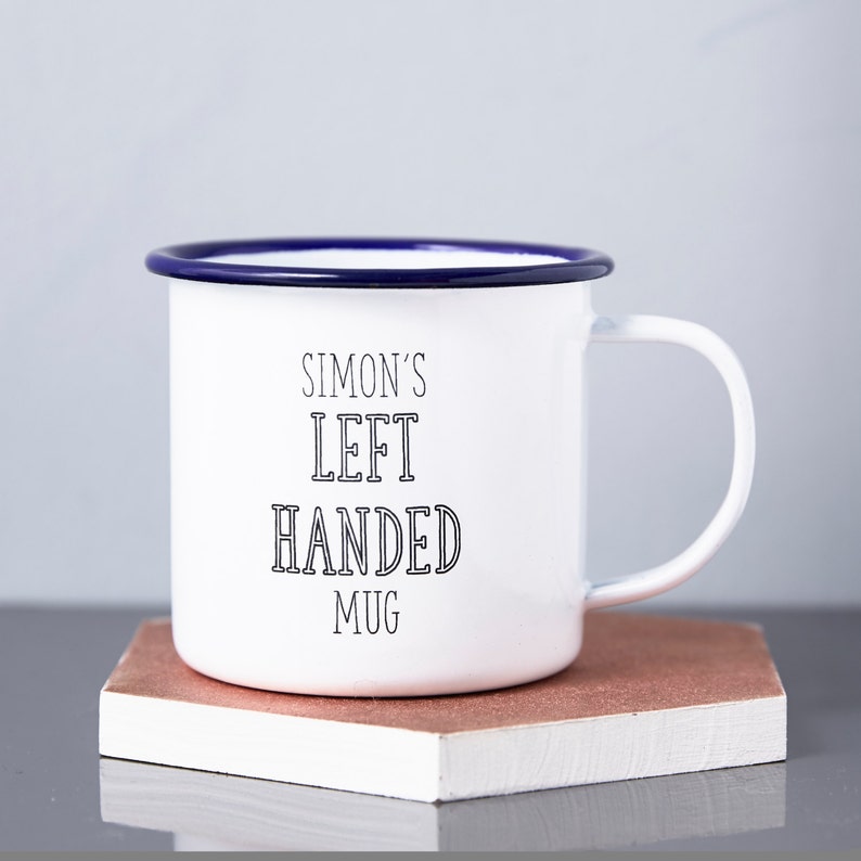 Może przedstawiać: Biały emaliowany kubek z niebieską obw&oacute;dką i uchwytem. Na kubku czarnym drukiem napisano "SIMON'S LEFT HANDED MUG". Kubek stoi na małym, prostokątnym, brązowym bloku.