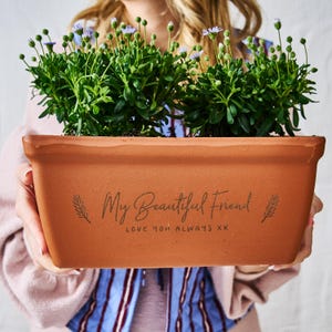 Può includere: Una fioriera rettangolare in terracotta piena di piante verdi e piccoli fiori viola. La fioriera ha l'iscrizione "My Beautiful Friend, Love You Always XX" in uno stile scritto a mano.