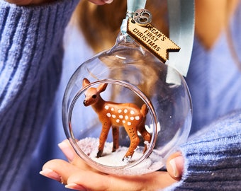 May include: A glass Christmas ornament with a small deer figurine inside. The ornament is filled with white glitter and has a wooden tag attached that says "Oscar's First Christmas".