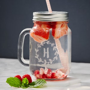 May include: A clear glass mason jar with a silver lid and a handle. The jar is filled with water, strawberries, and pomegranate seeds. A pink and white striped straw is in the jar. The jar has the letter "H" etched into it.