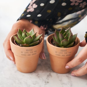 May include: Two terracotta pots with succulents, each pot has a personalized message. The pot on the left says "Simon My Love For You Grows And Grows". The pot on the right says "Jenny My Love For You Grows And Grows".