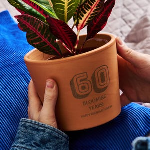 以下が含まれることがあります： テラコッタ色の植木鉢。数字の「60」と「Blooming Years! Happy Birthday Simon!」の文字がプリントされています。緑、赤、黄色の葉を持つ植物が入っています。2つの手で持っています。