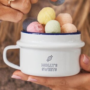 Pode incluir: Caneca de esmalte branco com borda azul e um logotipo de doce azul e branco. A caneca diz "Molly's Sweets" e está cheia de bolas de doce coloridas.
