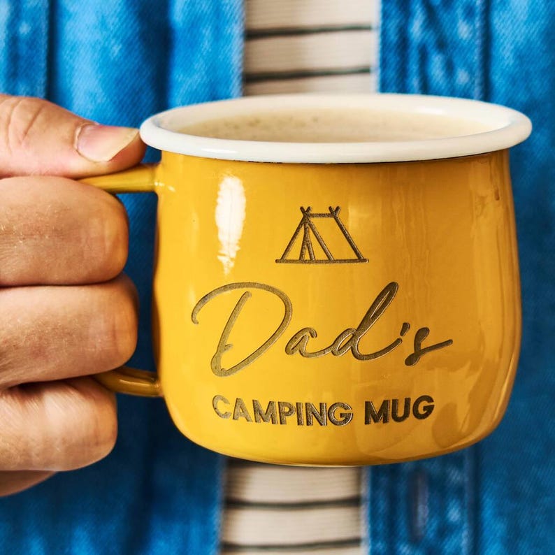 May include: A yellow camping mug with a white rim, filled with a light-colored beverage. The mug has the words "Dad's CAMPING MUG" etched into the side, along with a tent graphic. The mug is held by a person.