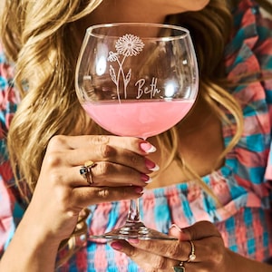Könnte beinhalten: Ein klares Weinglas mit einer rosa Flüssigkeit. Das Glas ist mit einem Blumendesign und dem Namen "Beth" graviert. Das Glas wird von einer Person gehalten, die Ringe und ein farbenfrohes Oberteil trägt.