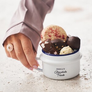 Pode incluir: Uma caneca de esmalte branco com uma borda azul e o texto "JESSICA'S Chocolate Treats" impresso no lado. A caneca está cheia de uma variedade de guloseimas de chocolate, incluindo uma barra de chocolate branco, uma trufa de chocolate, um quadrado de chocolate branco e um biscoito com granulado vermelho.