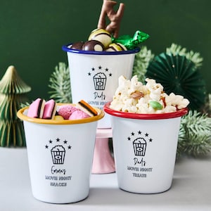 Pode incluir: Três canecas de esmalte branco com bordas coloridas, cheias de lanches. As canecas têm texto preto e um gráfico de pipoca. Uma caneca diz "Emma's Movie Night Snacks", outra diz "Dad's Movie Night Treats". A terceira caneca diz "Dad's Movie Night".