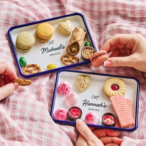 May include: Two white enamel trays with blue trim, each filled with a variety of snacks. One tray has the text "Michael's Snacks" and the other has the text "Hannah's Snacks".