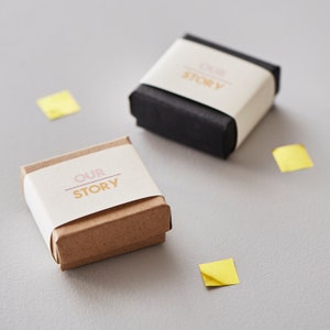 May include: Two small gift boxes, one black and one brown, with white bands printed with the words "OUR STORY" in pink and gold. Scattered around are small yellow square confetti pieces. The boxes are on a gray surface.