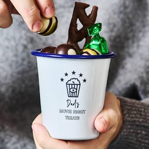 Pode incluir: Caneca esmaltada branca com borda azul, cheia de chocolates e doces. A caneca apresenta um gráfico de pipoca e o texto "Dad's Movie Night Treats". Uma mão está colocando um chocolate na caneca.