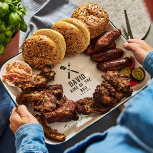 May include: A white rectangular platter with a blue trim, filled with barbecue food. The platter includes sesame seed buns, grilled sausages, ribs, coleslaw, and grilled vegetables. The platter has the text "DAVID KING OF THE BBQ".
