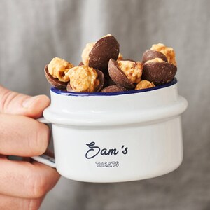 Pode incluir: Uma caneca de esmalte branco com uma borda azul e o texto "Sam's Treats" impresso na frente. A caneca está cheia de pipoca coberta de chocolate.