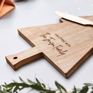 Pode incluir: Uma t&aacute;bua de servir de madeira castanho-clara em forma de &aacute;rvore de Natal com uma pega com um orif&iacute;cio. A t&aacute;bua est&aacute; gravada com o texto CHRISTMAS WITH The Jenson family. Uma faca de metal em tom de ouro rosa repousa na borda superior.