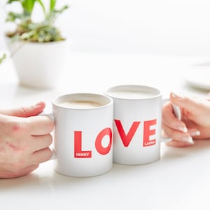 May include: Two white ceramic mugs with the word "LOVE" in red. One mug has "HENRY" and the other has "LAURA" printed below the word. Both mugs are filled with a light-coloured beverage and held by hands.