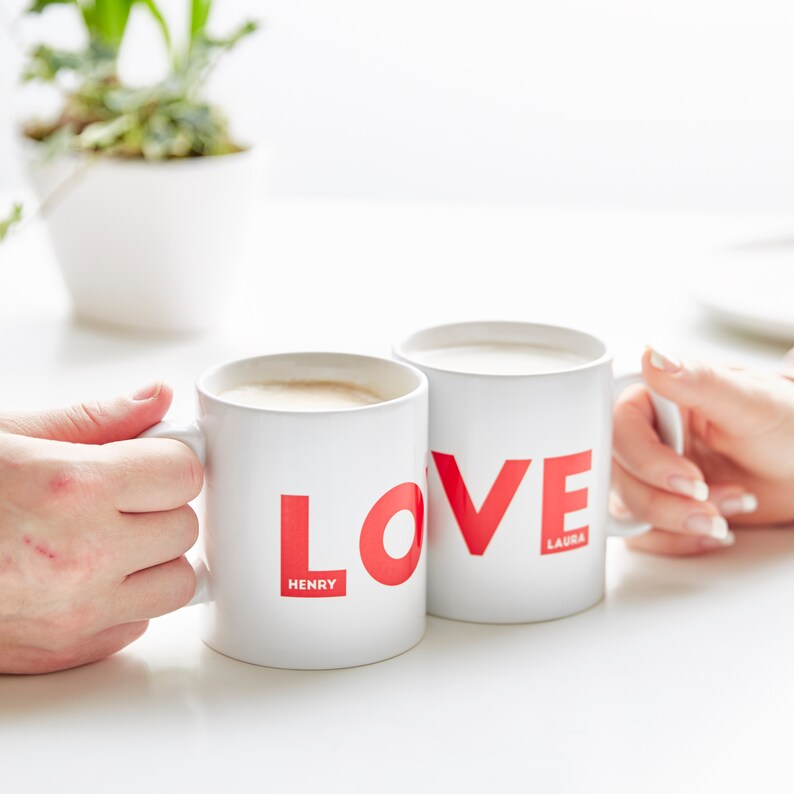 May include: Two white ceramic mugs with the word "LOVE" in red. One mug has "HENRY" and the other has "LAURA" printed below the word. Both mugs are filled with a light-coloured beverage and held by hands.