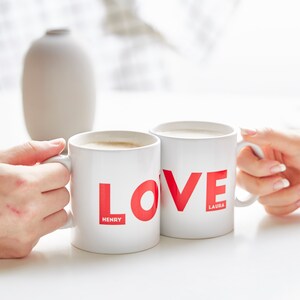 May include: Two white ceramic mugs with the word "LOVE" in red. The mug on the left has "HENRY" in a smaller red font below the "L", and the mug on the right has "LAURA" below the "E". Both mugs are filled with a light-coloured beverage.