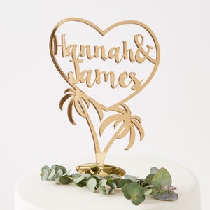 May include: Gold mirrored heart-shaped cake topper with the names "Hannah & James" inside the heart and two palm trees below.